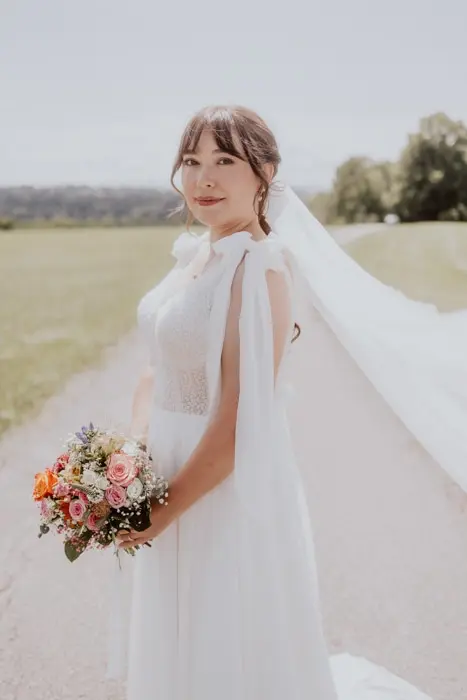 Hochzeitsfotograf Bayern Braut im weißen Kleid mit Blumenstrauß steht auf einem ländlichen Weg Dokumentarische Editorial Hochzeitsfotografie