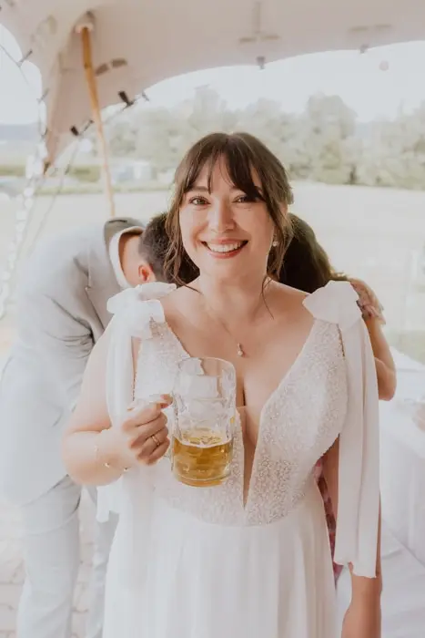 Hochzeitsfotograf Bayern Braut in weißem Kleid mit Bierkrug bei Hochzeit unter Pavillon Dokumentarische Editorial Hochzeitsfotografie