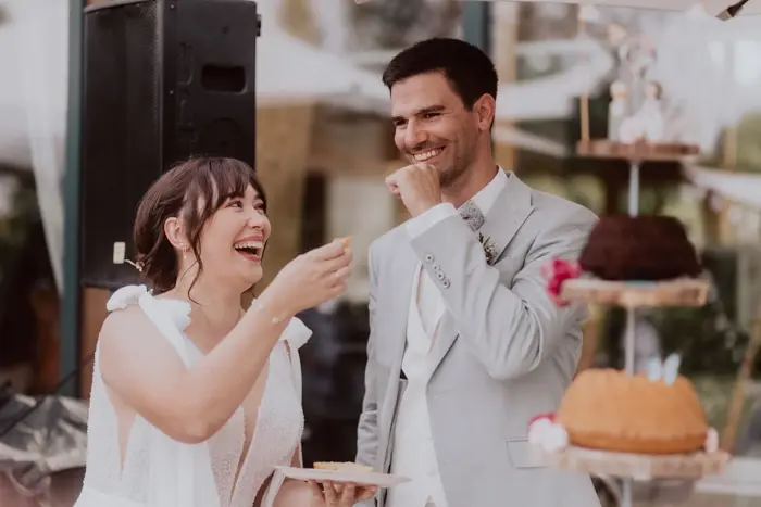 Hochzeitsfotograf Bayern Fröhliches Paar teilt Hochzeitstorte bei Empfang Dokumentarische Editorial Hochzeitsfotografie