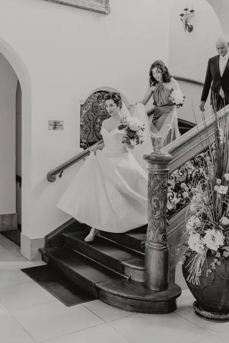 Hochzeitsfotograf Bayern Braut in weißem Kleid geht die Treppe hinunter, begleitet von Eltern Dokumentarische Editorial Hochzeitsfotografie