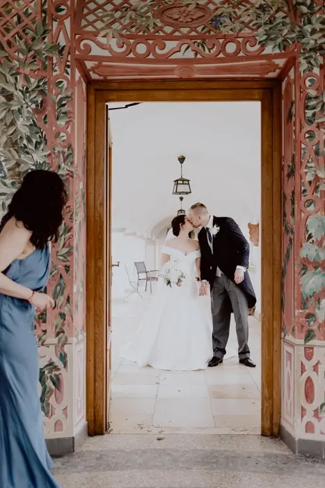 Hochzeitsfotograf Bayern Brautpaar küsst sich in Türrahmen, während Frau im blauen Kleid zuschaut Dokumentarische Editorial Hochzeitsfotografie