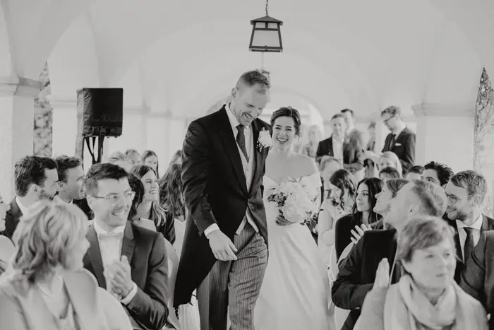 Hochzeitsfotograf Bayern Braut und Bräutigam gehen den Gang hinunter, umgeben von Gästen in einer Hochzeitszeremonie Dokumentarische Editorial Hochzeitsfotografie