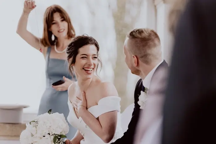 Hochzeitsfotograf Bayern Glückliches Brautpaar mit lachendem Gast im Hintergrund bei Hochzeit Dokumentarische Editorial Hochzeitsfotografie