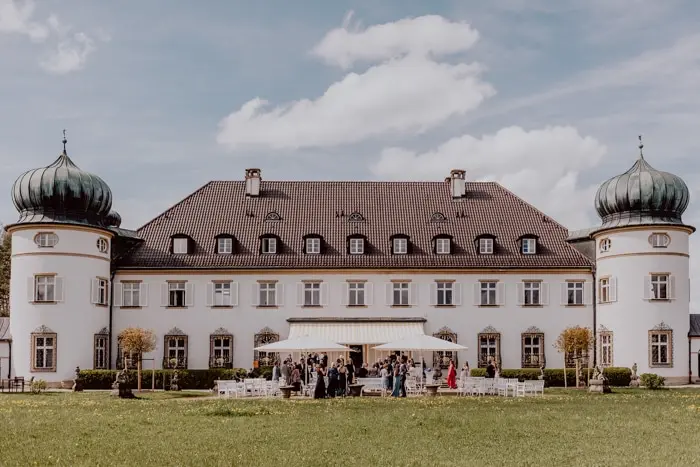 Hochzeitsfotograf Bayern Historisches Schloss mit Zwiebeltürmen und Personen auf dem Rasen Dokumentarische Editorial Hochzeitsfotografie