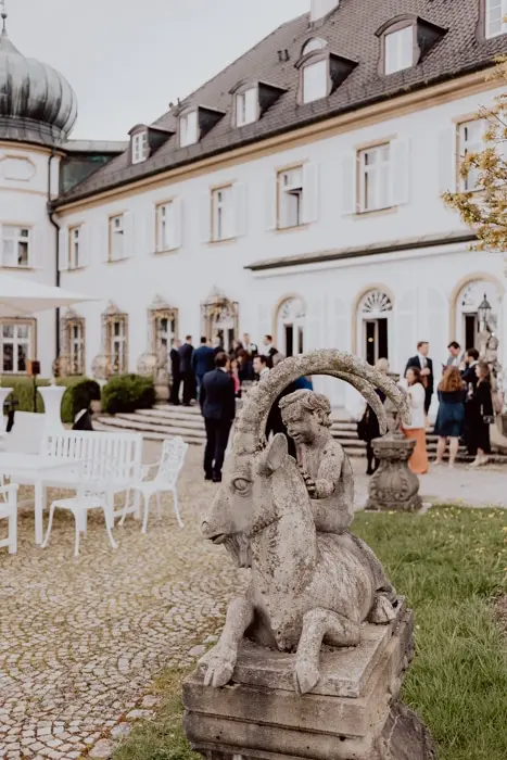 Hochzeitsfotograf Bayern Hochzeitsfeier vor einem historischen Gebäude mit Skulptur im Vordergrund Dokumentarische Editorial Hochzeitsfotografie