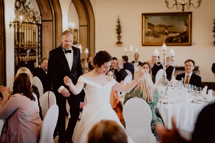 Hochzeitsfotograf Bayern Elegantes Hochzeitspaar betritt stilvoll dekorierten Festsaal Dokumentarische Editorial Hochzeitsfotografie