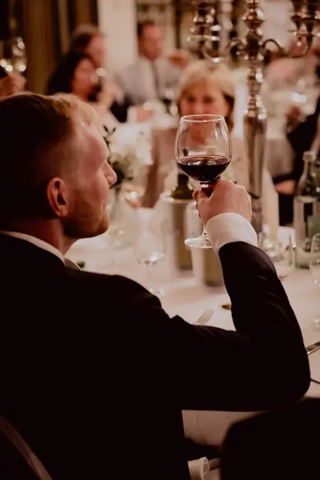 Hochzeitsfotograf Bayern Eleganter Mann in Anzug mit Rotweinglas bei festlichem Abendessen Dokumentarische Editorial Hochzeitsfotografie
