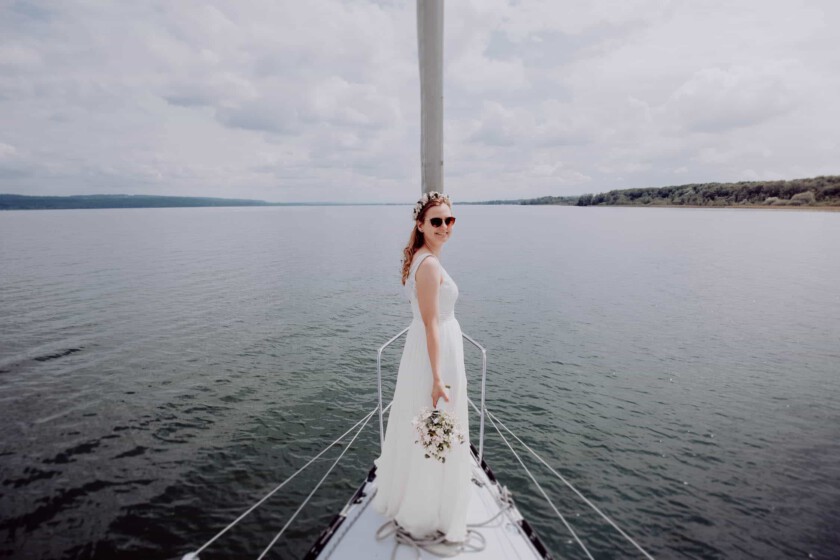 Braut steht vorne auf einem Segelboot und schaut Richtung Kamera. Sie trägt eine Sonnenbrille und hat einen Brautstrauß in der Hand. Im Hintergrund sieht man den Ammersee. Aufgenommen wurde das Bild vom Hochzeitsfotograf Ammersee