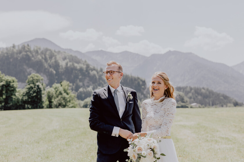 Brautpaar steht zusammen und lacht vor Bergkulisse auf der Hochzeit auf Gut Kaltenbrunn Tegernsee
