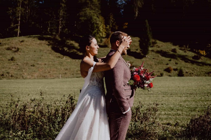 Braut hält dem Bräutigam von hinten die Augen zu beim First Look in Geitau. Im Hintergrund sieht man eine Wiese und Bäume.