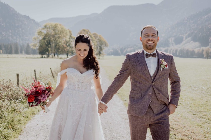 Braut und Bräutigam gehen lachend einen Weg entlang. Im Hintergrund sieht man eine helle alpine Landschaft.