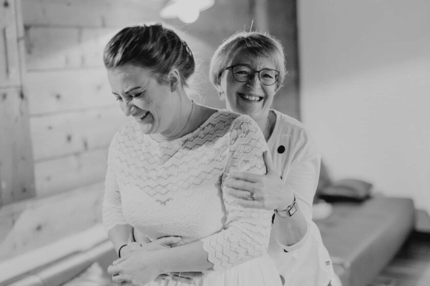 Braut und Brautmutter umarmen sich und lachen. Schwarzweiß Fotoaufnahme vom Hochzeitsfotograf Almbad Huberspitz.