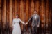Braut und Bräutigam stehen vor einer Holzwand, halten sich die Hände und lachen dem Hochzeitsfotograf in die Kamera. Hochzeit fand auf der Huberspitzalm statt.