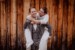 Bräutigam nimmt Braut Huckepack beim Brautpaarshooting auf der Huberspitzalm. Sie stehen vor einer Holzwand und Bräutigam schaut lachend in die Kamera
