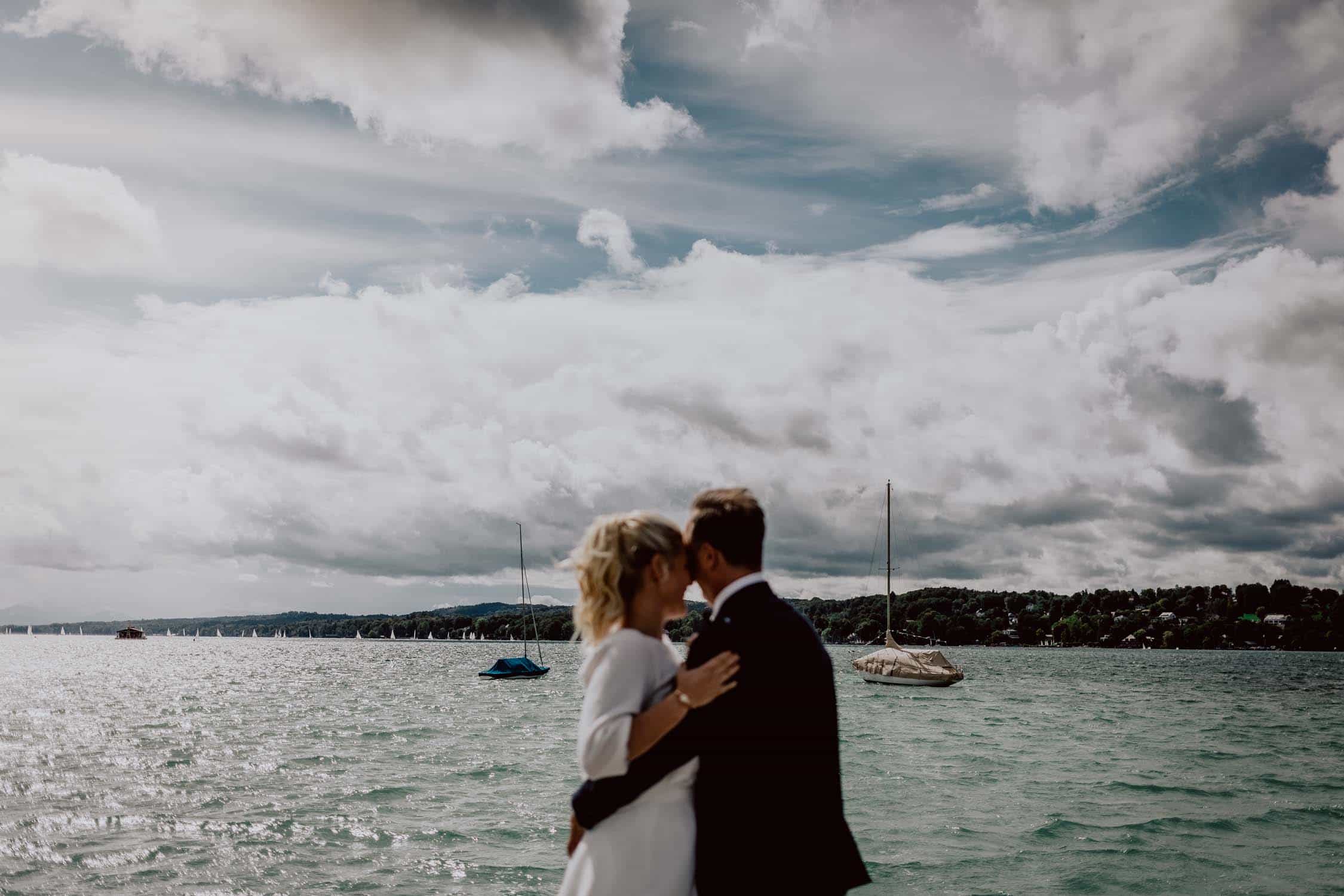 Brautpaar umarmt sich. Sie stehen vor dem Starnberger See. Man sieht Segelboote und einen teilweise bewölkten Himmel. Die Schärfe des Bildes liegt auf dem Hintergrund