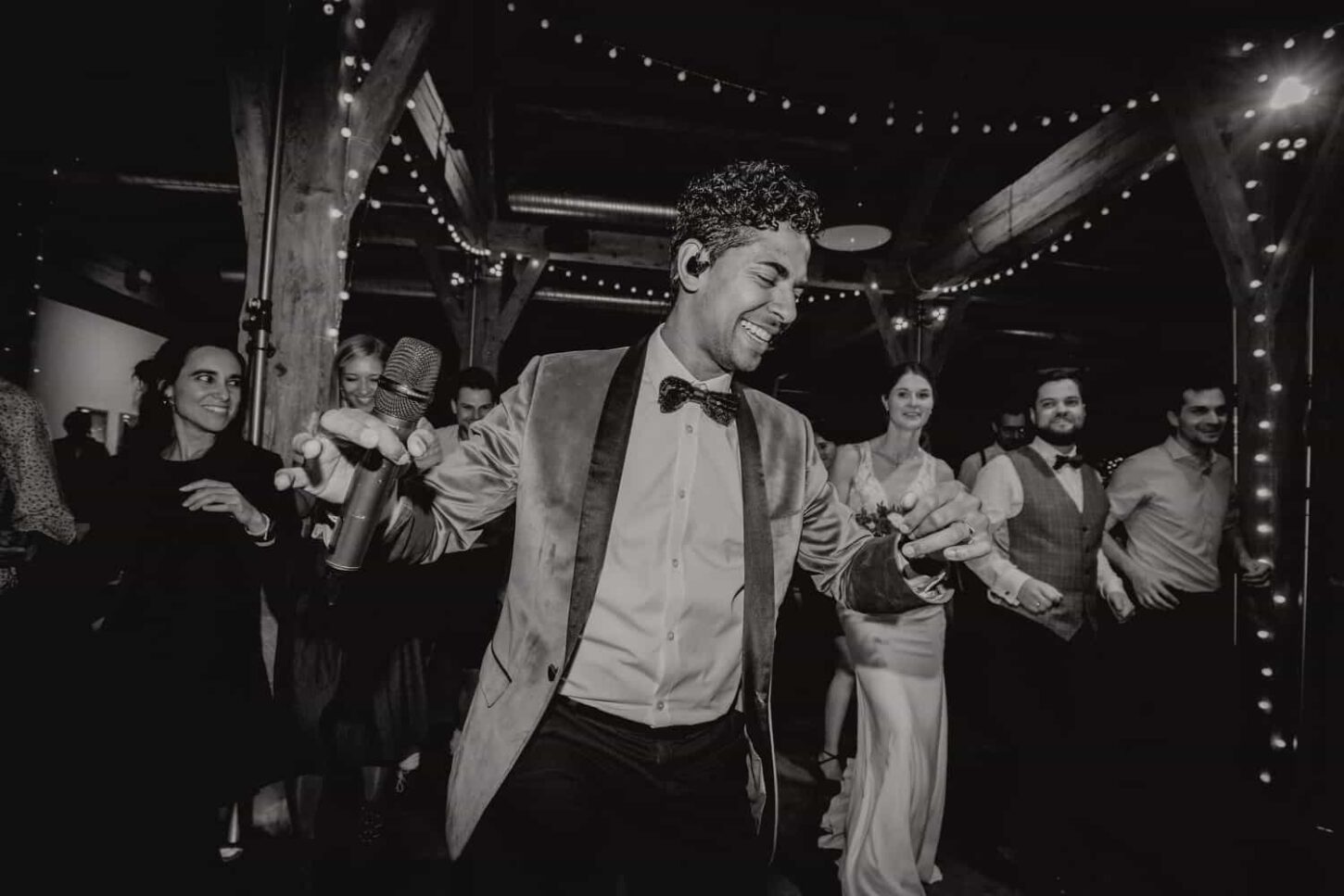 Hochzeitssänger lacht während seiner Performance. Im Hintergrund sieht man verschwommen das Brautpaar lachen. Es ist ein schwarz Weiß Foto.