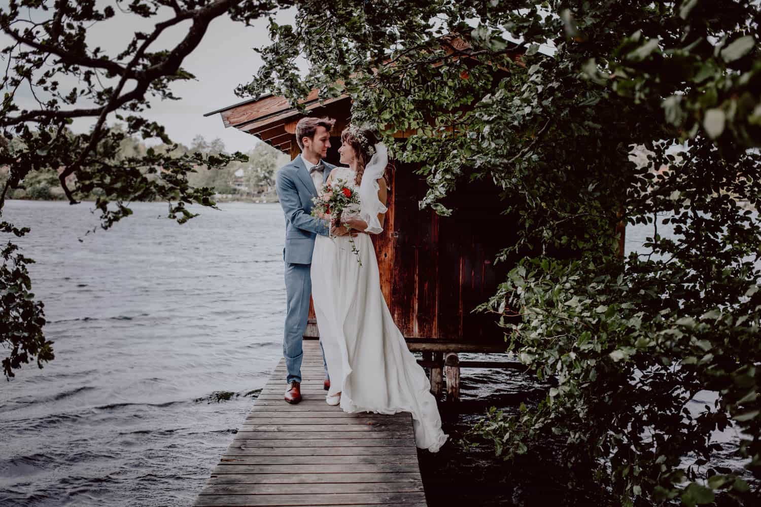 Brautpaar steht während ihrer Hochzeit im Fünfseeland auf einem Steg. Im Hintergrund sieht man den See und ein Bootshaus. Im Vordergrund Blätter und Zweige eines Baums.