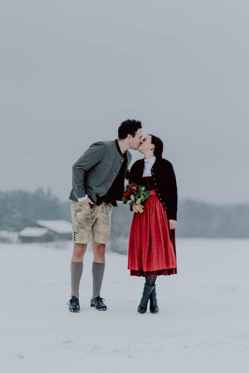Ein Paar geben sich einen Kuss während des Paarshooting. Sie stehen in einer verschneiten Landschaft und haben Dirndl und Lederhose an.