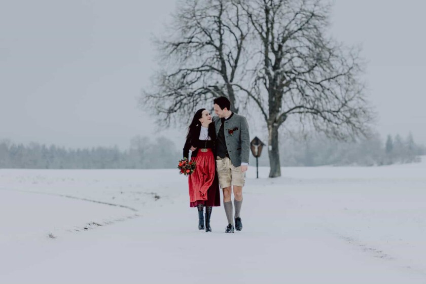 Ein Paar gehen Hand in Hand während des Paarshooting. Sie gehen in einer verschneiten Landschaft und haben Dirndl und Lederhose an.
