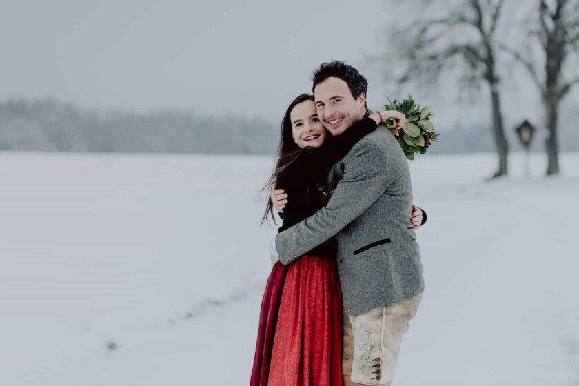 Ein Paar umarmt sich während des Paarshootings. Sie stehen in einer verschneiten Landschaft und haben Dirndl und Lederhose an.