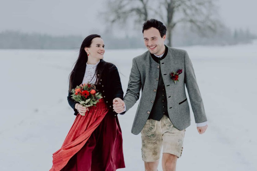 Ein Paar gehen Hand in Hand während des Paarshooting. Sie gehen in einer verschneiten Landschaft und haben Dirndl und Lederhose an.