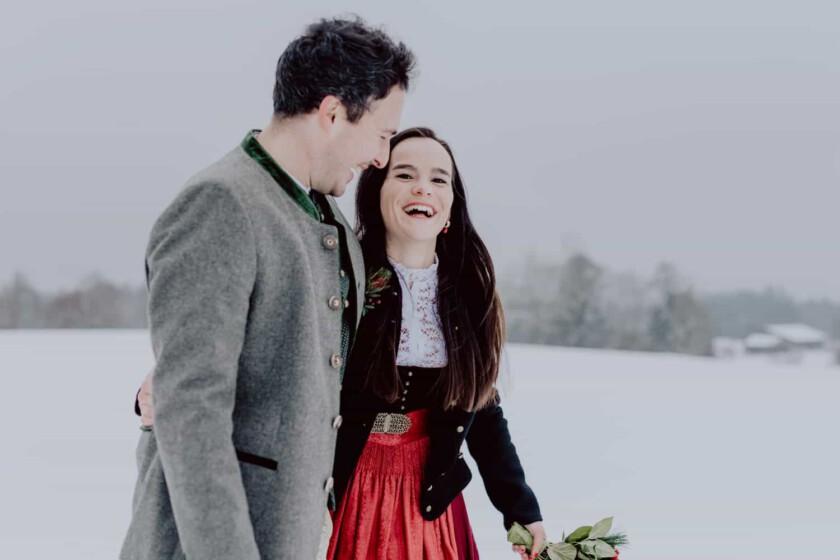 Ein Paar umarmt sich während des Paarshootings. Sie gehen in einer verschneiten Landschaft und haben Dirndl und Lederhose an.