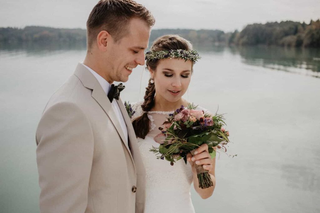 hochzeitsfotograf vintage - Brautpaar steht vor einem See. Die Braut schaut auf ihren Brautstrauß