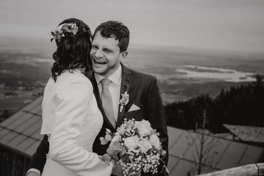 Schwarz Weiß Foto eines Brautpaares. Der Bräutigam lacht und umarmt die Braut. Das Hochzeitsfoto ist während der Hochzeit auf dem Hörnle in Bad Kohlgrub entstanden.