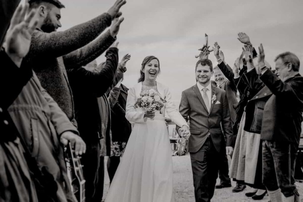 Schwarz Weiß Foto eines Brautpaares, das durch einen Spalier von Hochzeitsgästen läuft. Das Hochzeitsfoto ist während der Hochzeit auf dem Hörnle in Bad Kohlgrub entstanden.