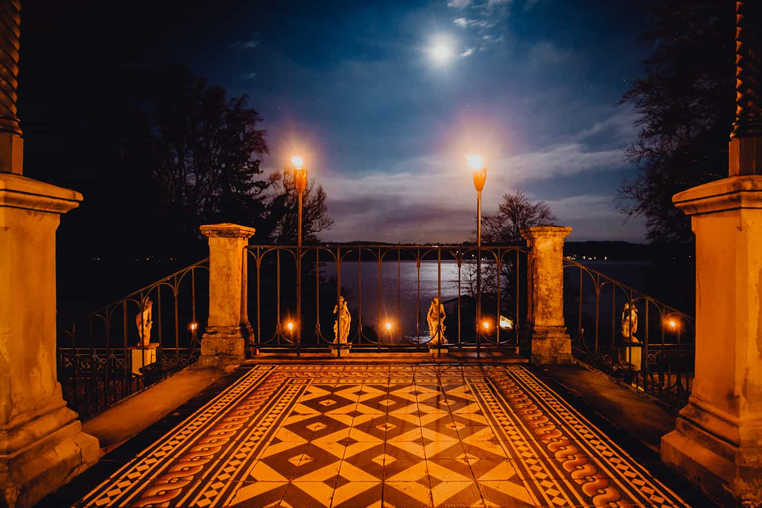 Nachtaufnahme der Terrasse La Villa am Starnberger See. Man sieht Fackeln, die den Boden orange beleuchten und im Himmel den Mond, der den Starnberger See blau beleuchtet