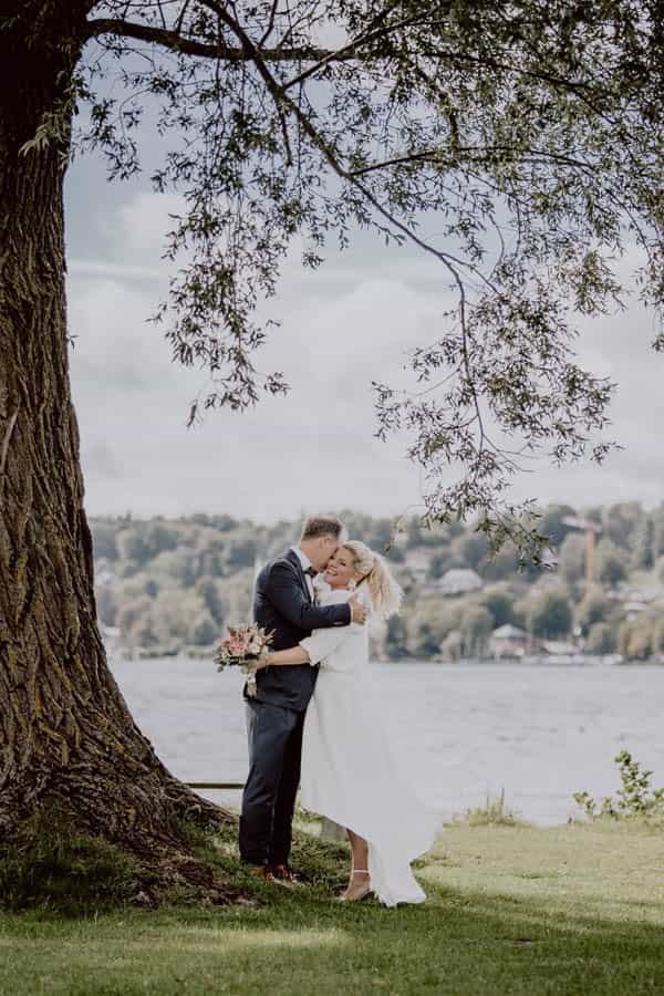 Hochzeitsfotograf Starnberger See - Aufnahme eines Brautpaares vor der See. Sie umarmen sich und stehen neben einem Baum. Im Hintergrund sieht man den Starnberger See