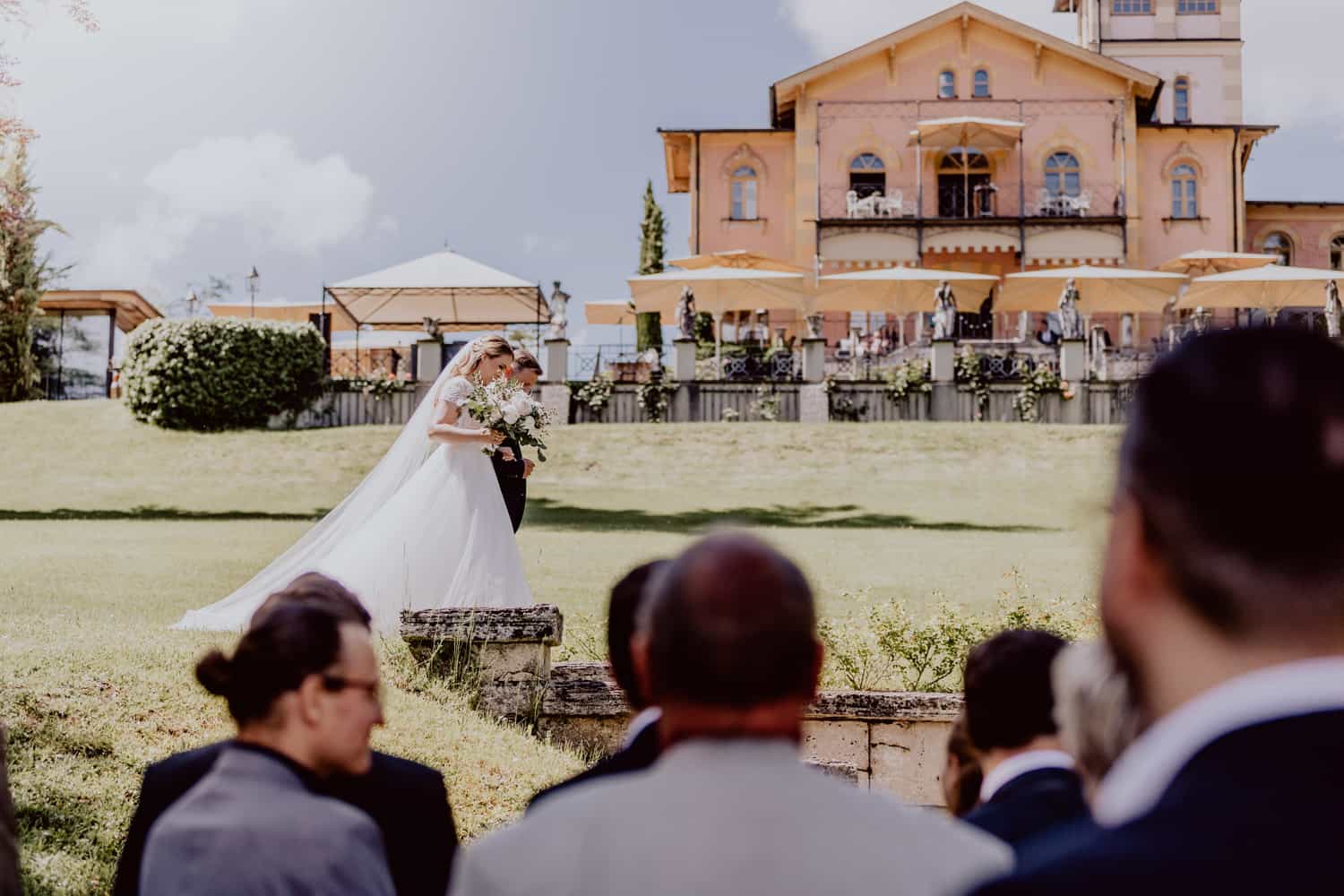 Braut geht mit Brautvater zur Trauung. Man sieht im Hintergrund die La Villa am Starnberger See. Im Vordergrund sieht man Hochzeitsgäste, die zur Braut schauen. Es ist ein sonniger Tag mit Blauem Himmel. Das Foto wurde vom Hochzeitsfotograf La Villa in Farbe aufgenommen.