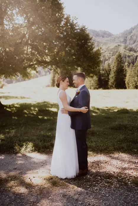 Hochzeitsfotograf Bayern Brautpaar umarmt sich im sonnigen Park vor grüner Landschaft Dokumentarische Editorial Hochzeitsfotografie