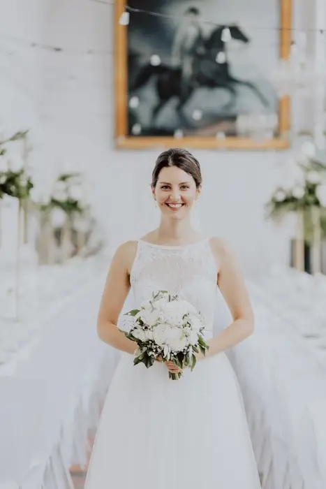 Hochzeitsfotograf Bayern Braut mit Blumenstrauß in einem eleganten weißen Raum mit Pferdegemälde Dokumentarische Editorial Hochzeitsfotografie